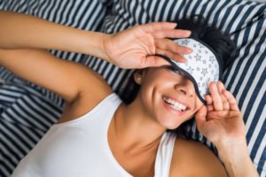 portrait young attractive brunette woman lying on bed in pajamas and sleeping mask, smiling in bedroom, happy emotion, lazy in morning, wake up, white teeth.