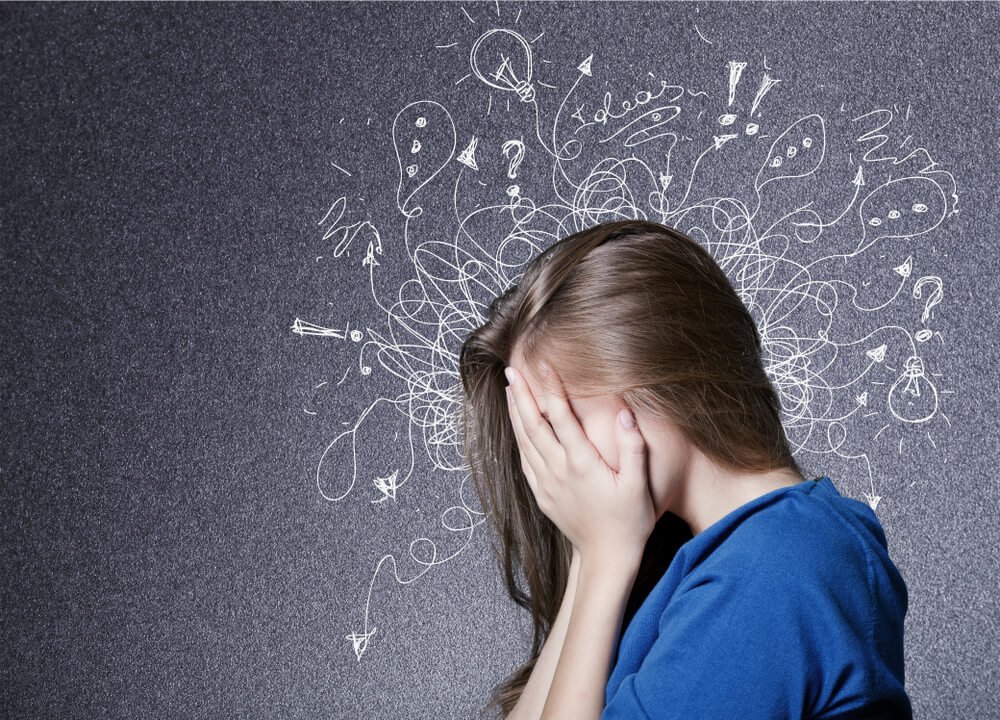 A woman holds her head in her hands; erratic lines and drawings circle her head, indicating anxiety.
