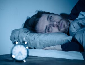 A man with a sleep disorder laying in a bed staring at an alarm clock.