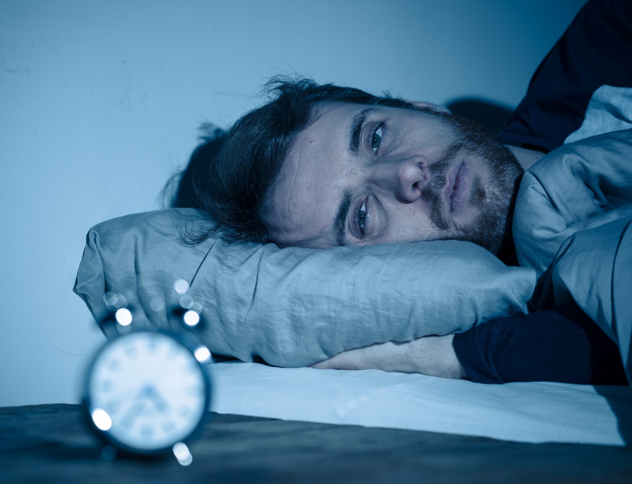 A man with a sleep disorder laying in a bed staring at an alarm clock.
