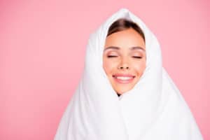 A woman is completely wrapped in a white wool comforter, set against a pastel pink background.