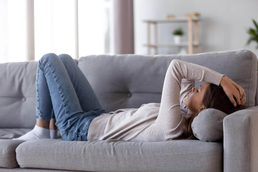 A woman in jeans and a gray shirt holding her head and laying down on a gray couch.