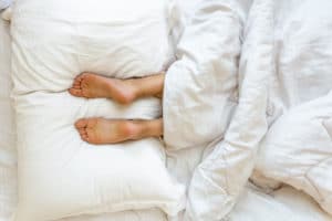 Two feet resting on two white bed pillows underneath white bed covers.