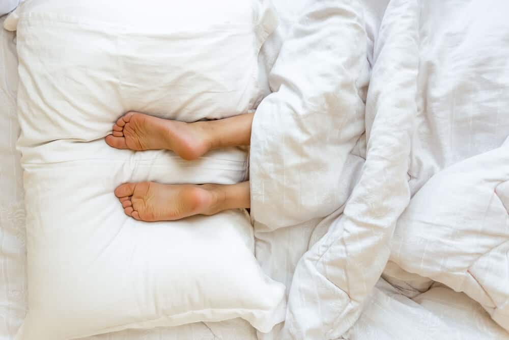 Two feet resting on two white bed pillows underneath white bed covers.