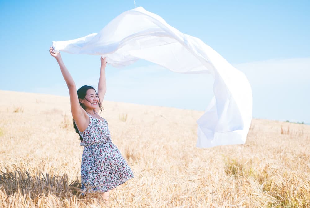 A woman holds a sheet above her head in a field on a sunny day.